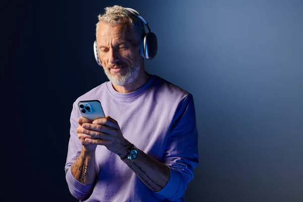 Alegre atractivo hombre maduro con barba y auriculares disfrutando de la música y mirando su teléfono - foto de stock