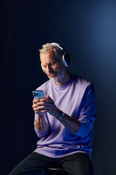 Good looking bearded mature man in purple sweatshirt with headphones looking at his smartphone — Stock Photo