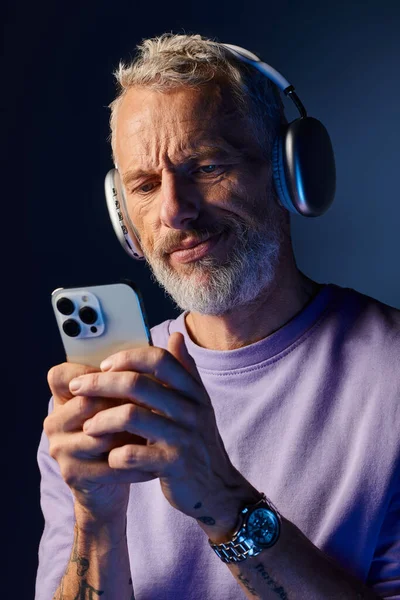 Hombre maduro barbudo de buen aspecto en sudadera púrpura con auriculares mirando su teléfono inteligente - foto de stock