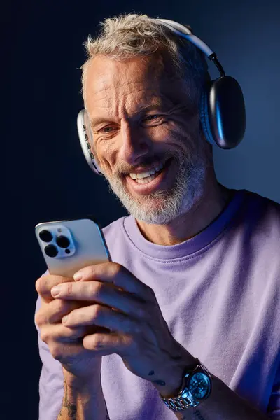 Веселий красивий зрілий чоловік з бородою і навушниками насолоджується музикою і дивиться на його телефон — стокове фото