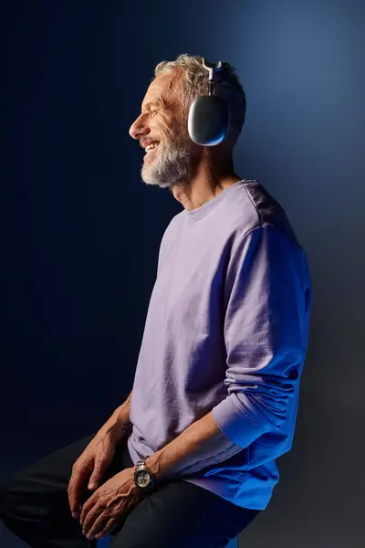 Joyful attractive mature man with gray beard and headphones in purple sweatshirt enjoying music — Stock Photo