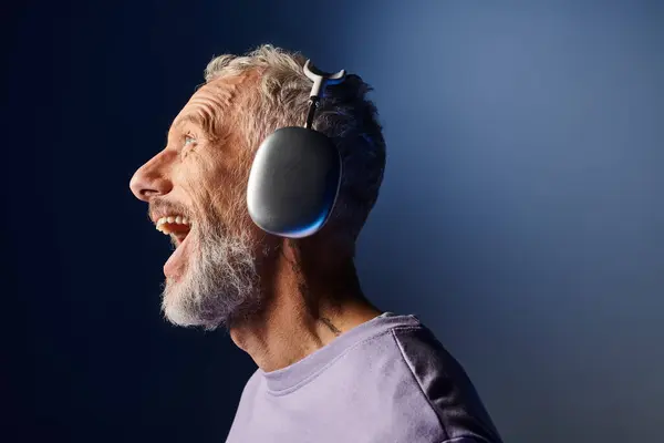 Cheerful handsome mature man with gray beard and headphones in purple sweatshirt enjoying music — Stock Photo