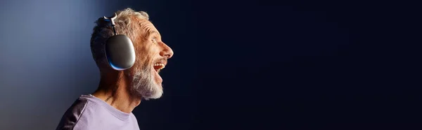 Cheerful handsome mature man with beard and headphones in purple sweatshirt enjoying music, banner — Stock Photo