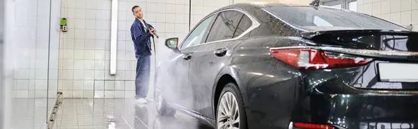 Attrayant spécialiste dévoué dans confortable uniforme bleu en utilisant un tuyau pour laver la voiture moderne noire, bannière — Photo de stock