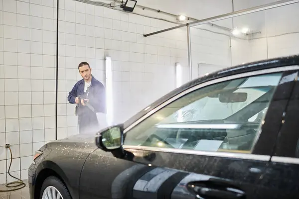 Attractive dedicated professional in blue uniform using hose to wash modern black car in garage — Stock Photo