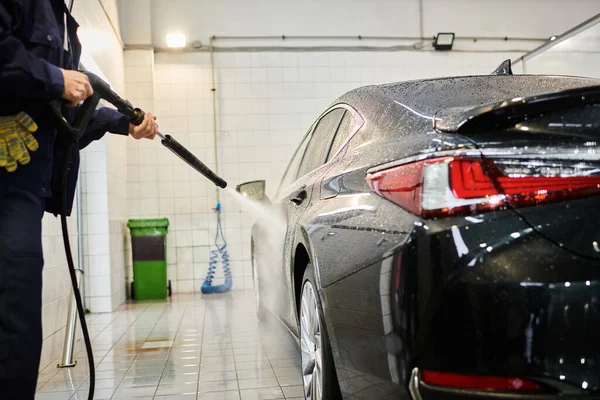 Cropped view of dedicated serviceman in blue uniform using hose to wash black car in garage — Stock Photo