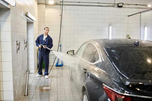 Good looking devoted serviceman in blue uniform with collected hair using hose to wash black car — Stock Photo