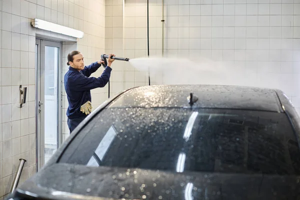 Bonito dedicado trabalhador profissional em uniforme usando mangueira para lavar carro moderno preto enquanto na garagem — Fotografia de Stock