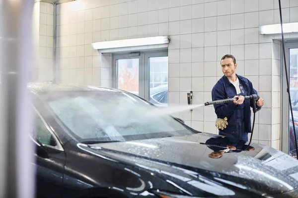 Bom olhar dedicado serviceman com cabelos recolhidos em uniforme lavagem de carro preto, enquanto na garagem — Fotografia de Stock