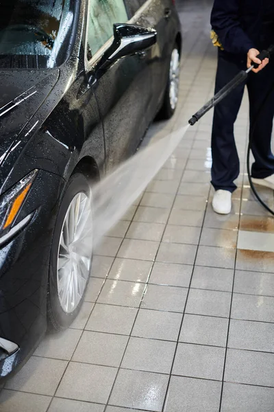 Обрізаний вид спеціального професійного працівника в блакитному кольорі задовольняє рівномірне миття чорного автомобіля в гаражі — стокове фото