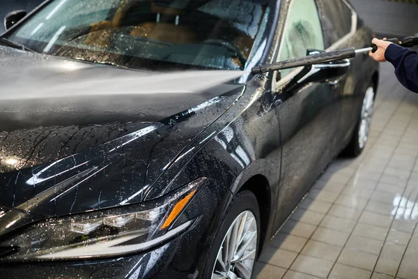 Cropped view of hard working specialist in blue uniform using hose to wash black modern car — Stock Photo