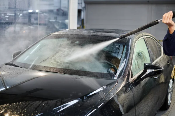 Cropped view of devoted professional serviceman in uniform washing black car while in garage — Stock Photo