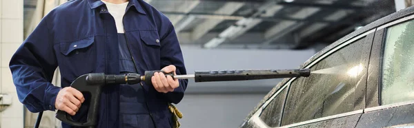 Cropped view of hard working professional in blue uniform using hose to wash black car, banner — Stock Photo
