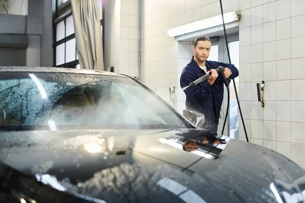 Good looking devoted specialist in comfy blue uniform with collected hair washing car in garage — Stock Photo