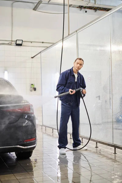 Attraktive, hart arbeitende Profis in blauer Uniform mit gesammelten Haaren, die mit einem Schlauch das Auto waschen — Stockfoto