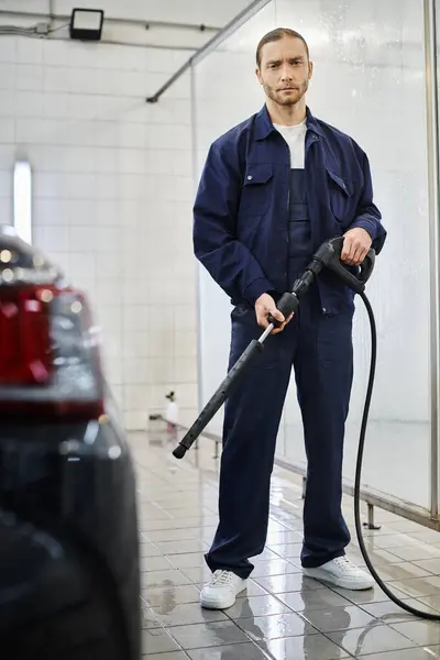 Schöner professioneller Servicemann in blauer Uniform, der einen Schlauch hält, bevor er ihn benutzt, um schwarzes Auto zu waschen — Stockfoto
