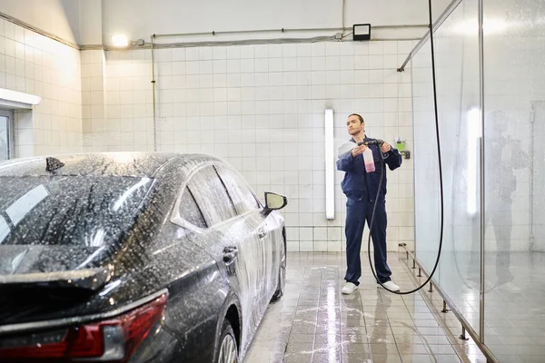 Handsome professional worker in blue uniform using soap to wash black modern car in garage — Stock Photo