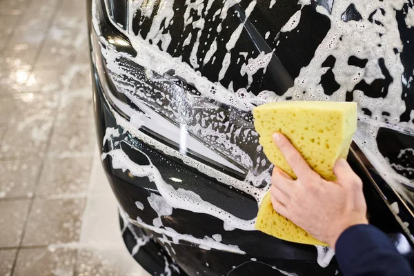 Cropped view of hard working professional in blue uniform washing car with soapy sponge in garage — Stock Photo