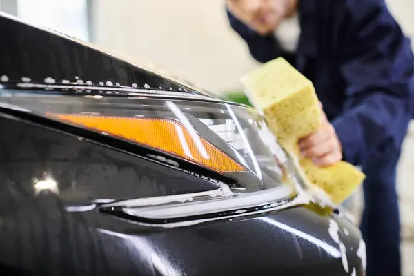 Se concentrer sur l'éponge savonneuse dans les mains d'un travailleur attrayant dédié flou lavage voiture noire dans le garage — Photo de stock