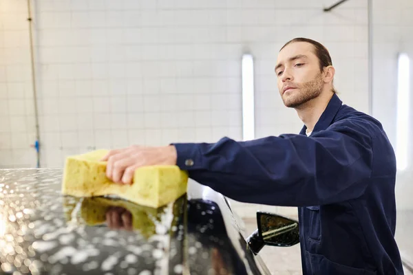 Atraente dedicado serviceman no confortável carro de lavagem uniforme azul com esponja ensaboada, enquanto na garagem — Fotografia de Stock