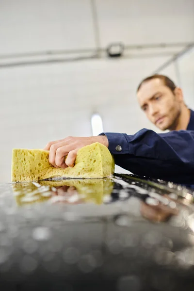 Focus on yellow soapy sponge in hands of blurred handsome man in uniform washing black car — Stock Photo