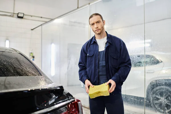 Handsome professional worker in blue uniform holding soapy sponge and looking at camera in garage — Stock Photo