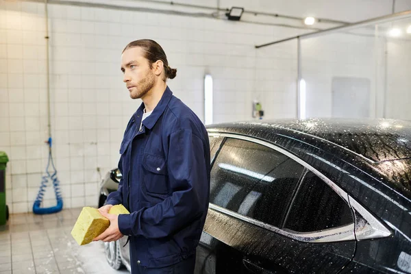Handsome professional serviceman in blue uniform with collected hair holding sponge next to car — Stock Photo