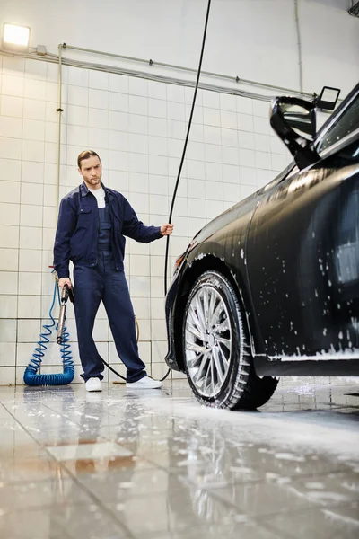 Attractive enthusiastic serviceman with collected hair in blue uniform washing modern car using hose — Stock Photo