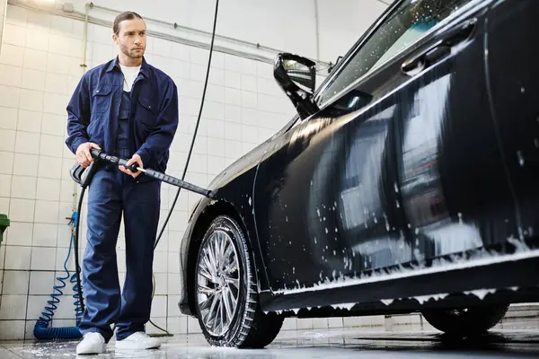 Appealing dedicated specialist in blue uniform with collected hair washing car with hose in garage — Stock Photo