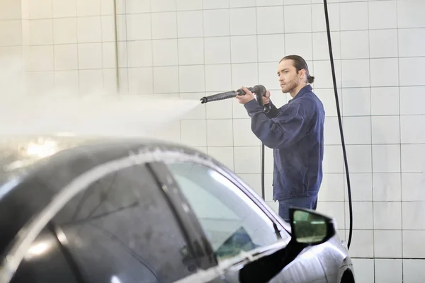 Handsome hard working serviceman in blue uniform with collected hair washing car with hose — Stock Photo