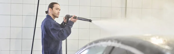 Dedicated handsome serviceman in blue uniform with collected hair washing car with hose, banner — Stock Photo