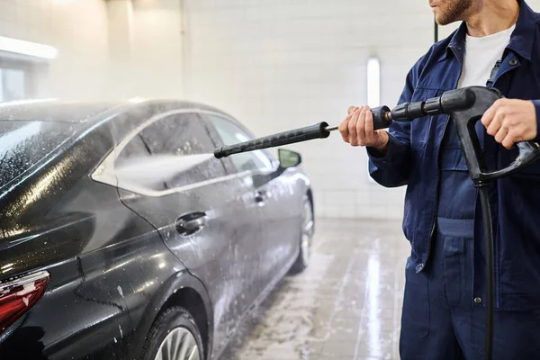 Cropped view of devoted professional worker in blue uniform using hose to wash black modern car — Stock Photo