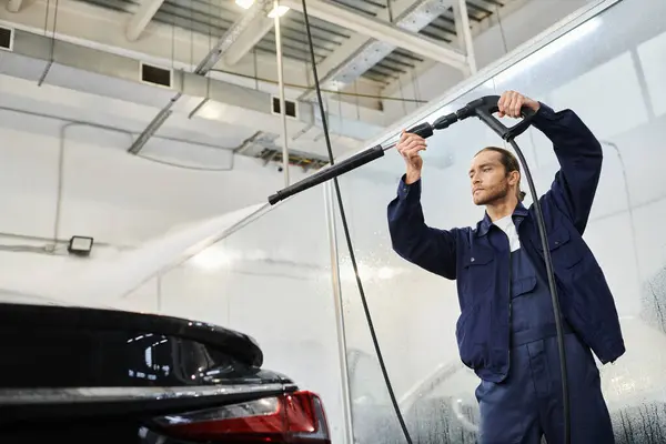 Schöner engagierter begeisterter Spezialist in blauer Uniform mit Schlauch, um schwarzes Auto in der Garage zu reinigen — Stockfoto
