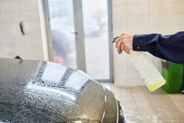 Cropped view of enthusiastic professional worker in uniform using pulverizer with water to clean car — Stock Photo