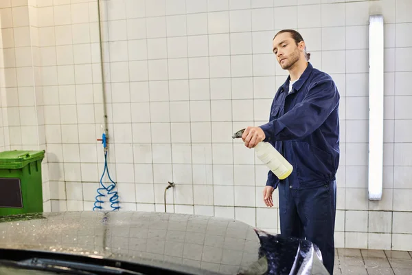 Добре виглядає важко працюючий чоловік зібраним волоссям у синій формі, використовуючи пульверизатор для очищення автомобіля — стокове фото