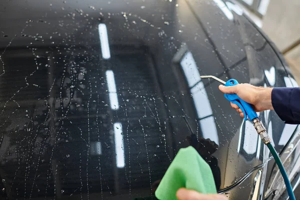 Cropped view of hard working professional in uniform using water hose and rag to clean black car — Stock Photo