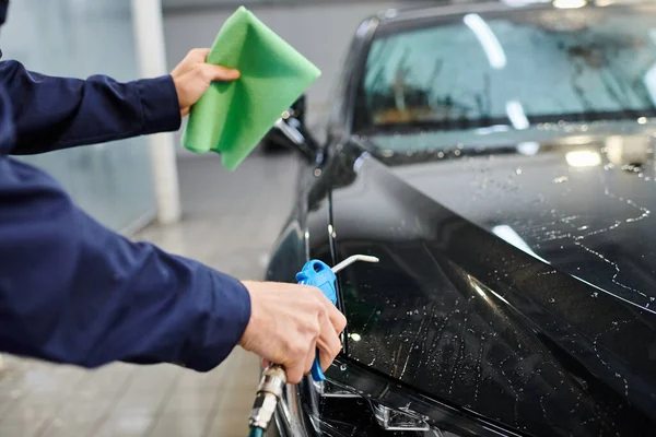Cropped view of dedicated professional in uniform using water hose and rag to clean modern black car — Stock Photo