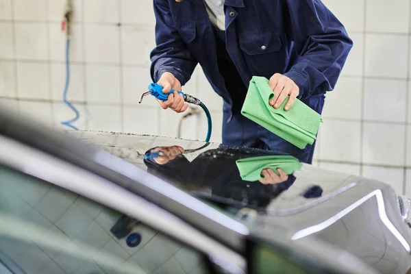 Cropped view of devoted serviceman in uniform using rag and hose to clean black car in garage — Stock Photo