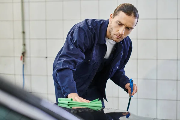 Schöner engagierter professioneller Servicemann mit gesammelten Haaren, der Lappen benutzt, um schwarzes Auto zu reinigen — Stockfoto