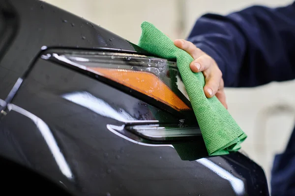 Visão cortada de especialista dedicado trabalho duro em uniforme usando pano para limpar carro moderno preto — Fotografia de Stock