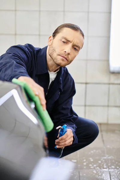 Appealing devoted serviceman in blue uniform with collected hair using hose and rag to clean car — Stock Photo