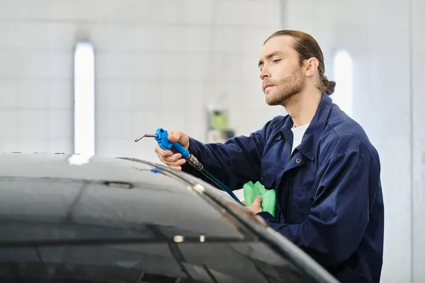 Atraente trabalhador duro serviceman em carro de limpeza uniforme azul com mangueira e pano verde na garagem — Fotografia de Stock