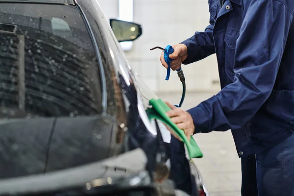 Cropped view of dedicated professional worker in uniform holding hose and cleaning car with rag — Stock Photo