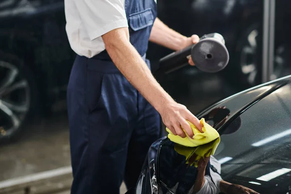 Cropped view of hard working serviceman cleaning black car with rag and holding polishing machine — Stock Photo