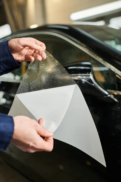 Visão recortada do especialista em trabalho duro em uniforme azul pronto para aplicar folha protetora no espelho do carro — Fotografia de Stock