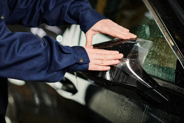 Cropped view of hard working man in uniform applying protective foil on side view mirror of car — Stock Photo