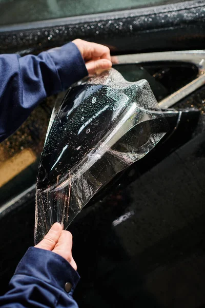 Cropped view of professional hard working serviceman in uniform applying protective foil on car — Stock Photo