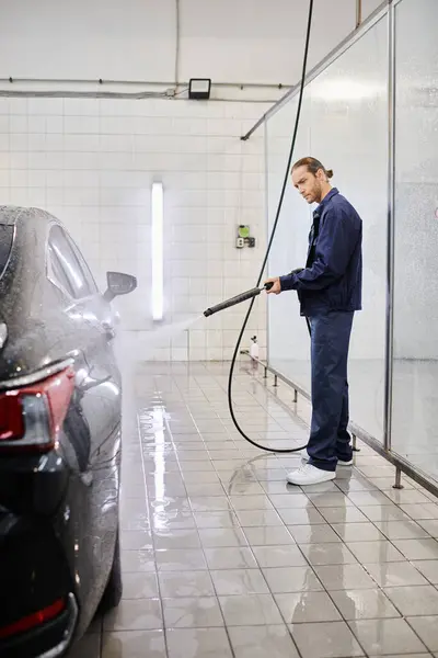 Appealing professional male worker in blue uniform washing black car with hose while in garage — Stock Photo