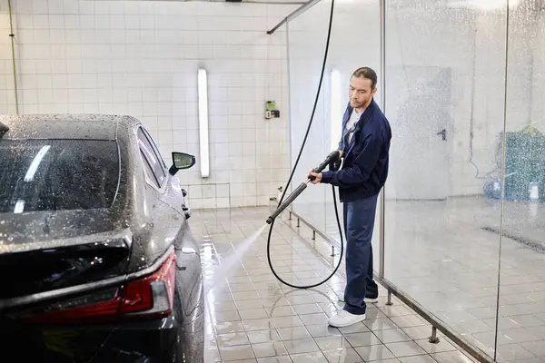 Good looking dedicated worker in blue uniform washing black car using hose while in garage — Stock Photo