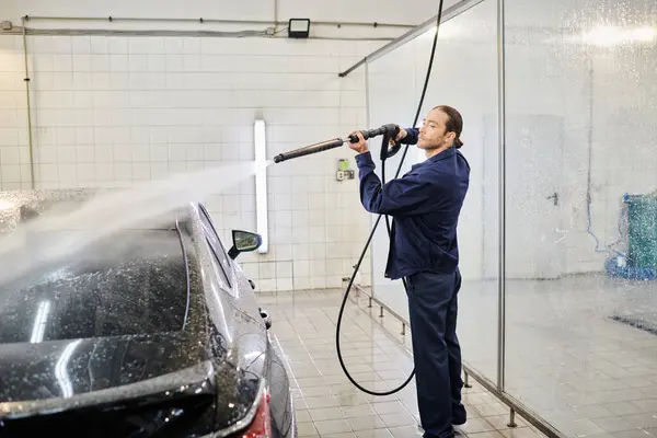 Attractive hard working man in blue uniform using hose to clean black modern car in garage — Stock Photo
